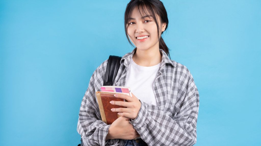 A young asian woman holding a book and a backpack.